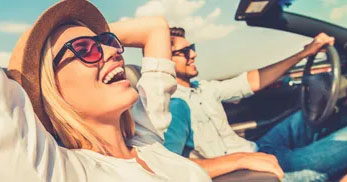 A couple relaxes in a convertible, enjoying a sunny day with a clear sky and distant scenery. The atmosphere is carefree and joyful.