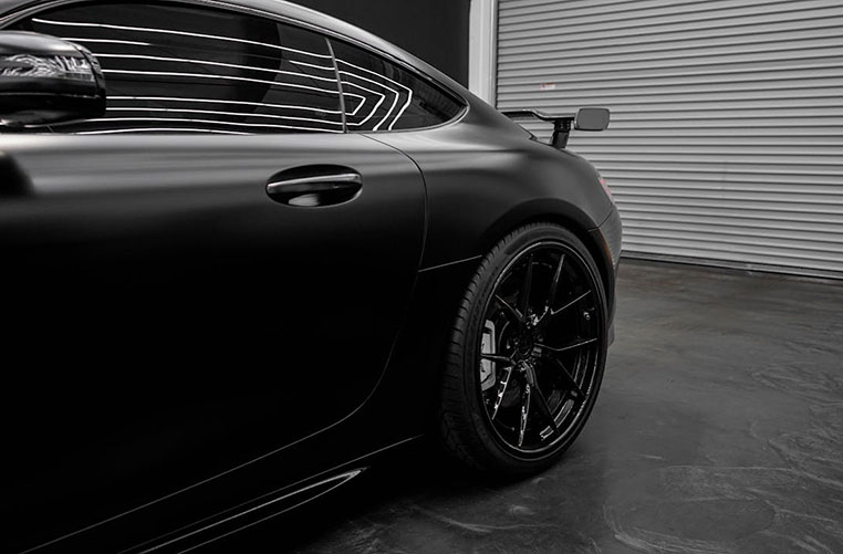 A sleek, matte black sports car with a large rear spoiler and black alloy wheels, showcased against a modern garage backdrop.