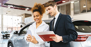 A car salesperson and a customer review documents in a modern car dealership, with a vehicle visible in the background.